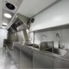The 3.8m Big Box Food Trailer features a modern commercial kitchen with stainless steel countertops, sinks, and cabinets. It includes an overhead ventilation system with a clean, industrial design and bright overhead lights illuminating the area.
