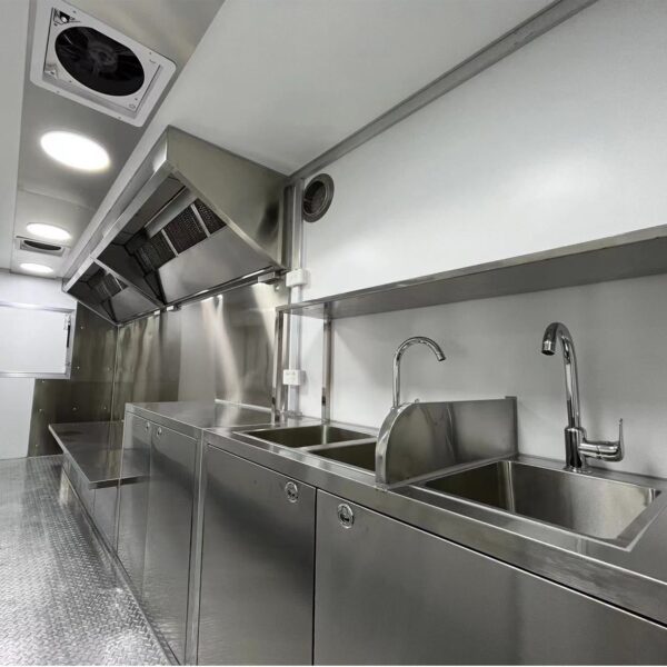 The 3.8m Big Box Food Trailer features a modern commercial kitchen with stainless steel countertops, sinks, and cabinets. It includes an overhead ventilation system with a clean, industrial design and bright overhead lights illuminating the area.