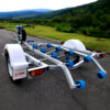 The Galvanised Single Jet-Ski Trailer ATM 750KG is shown on a paved road, featuring white fenders, blue rollers, and a spare tire mounted on the left side, with lush green hills and a cloudy sky in the background.