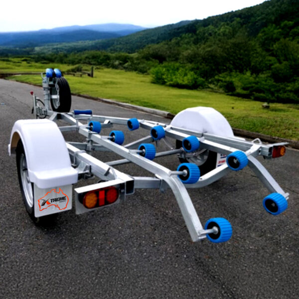 The Galvanised Single Jet-Ski Trailer ATM 750KG is shown on a paved road, featuring white fenders, blue rollers, and a spare tire mounted on the left side, with lush green hills and a cloudy sky in the background.