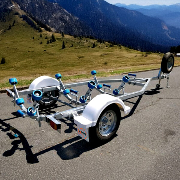 A Galvanised Single Jet-Ski Trailer ATM 750KG outfitted with blue rollers rests on a paved road, offering a view of green hills and distant peaks under a clear blue sky.