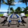 A Galvanised Single Jet-Ski Trailer ATM 750KG with blue rollers is parked on a sunny road lined with palm trees, set against an ocean and clear blue sky backdrop.