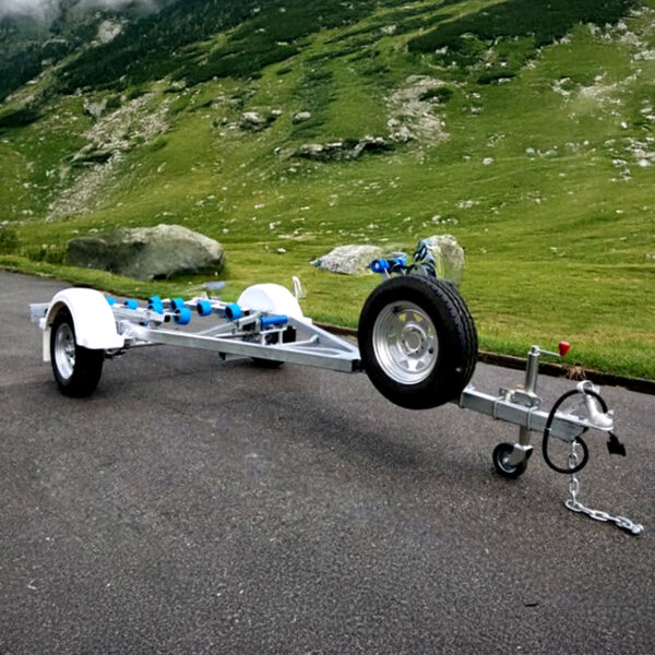 A Galvanised Single Jet-Ski Trailer ATM 750KG with a spare tire at the front is parked on an asphalt surface, set against a lush green landscape of rolling hills and scattered rocks beneath a cloudy sky.
