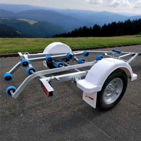A Galvanised Single Jet-Ski Trailer with blue rollers, two white wheel arches, and a metal frame is parked on a paved area near mountainous scenery with trees.