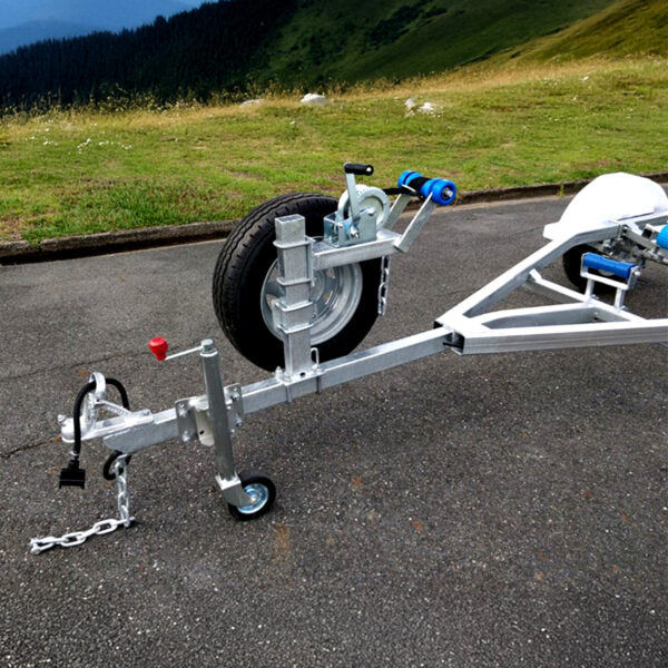 Close-up of the Galvanised Single Jet-Ski Trailer ATM 750KG with a mounted spare tire on a quiet road, featuring a silver frame, blue rollers, hand crank, small maneuvering wheel, and safety chain against a grassy hillside backdrop.