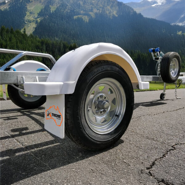 A close-up of a Galvanised Single Jet-Ski Trailer ATM 750KG wheel with a silver rim on a paved road. Another wheel and trailer hitch are visible, set against a mountainous landscape with lush green trees and snow-dusted peaks under a blue sky.