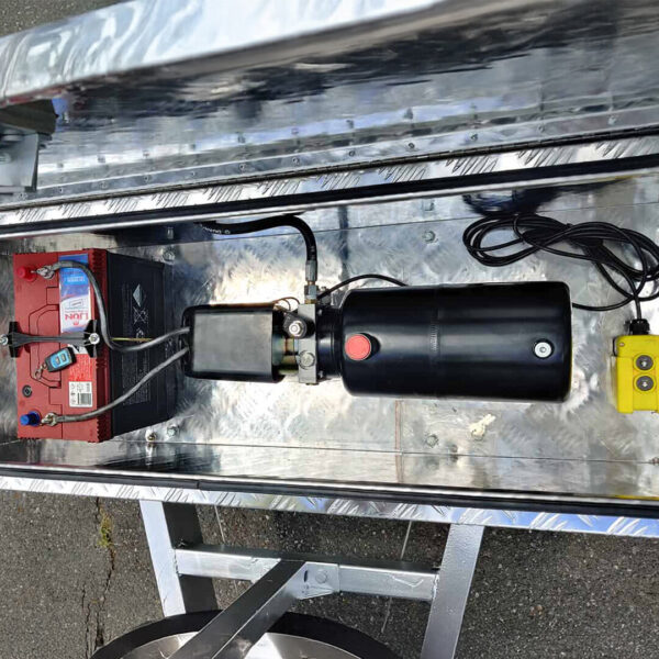 A close-up view of the 14x7 Dual Axle Hydraulic Tipper Tandem Box Trailer - 4500KG ATM With Ramps, showing its metal compartment with red battery, black hydraulic pump, wiring, and yellow control pendant on the trailer’s platform outdoors.
