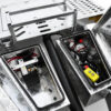 Open metal compartments of the Premium Hydraulic Tipper Car Trailer 4200kg ATM 16.4x6.6FT (5x2M) Deck Size – Punched Floor reveal electrical components, including a battery, cables, and a yellow control unit with buttons, all mounted on the trailer’s punched, diamond-patterned metal deck.