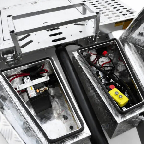 Open metal compartments of the Premium Hydraulic Tipper Car Trailer 4200kg ATM 16.4x6.6FT (5x2M) Deck Size – Punched Floor reveal electrical components, including a battery, cables, and a yellow control unit with buttons, all mounted on the trailer’s punched, diamond-patterned metal deck.